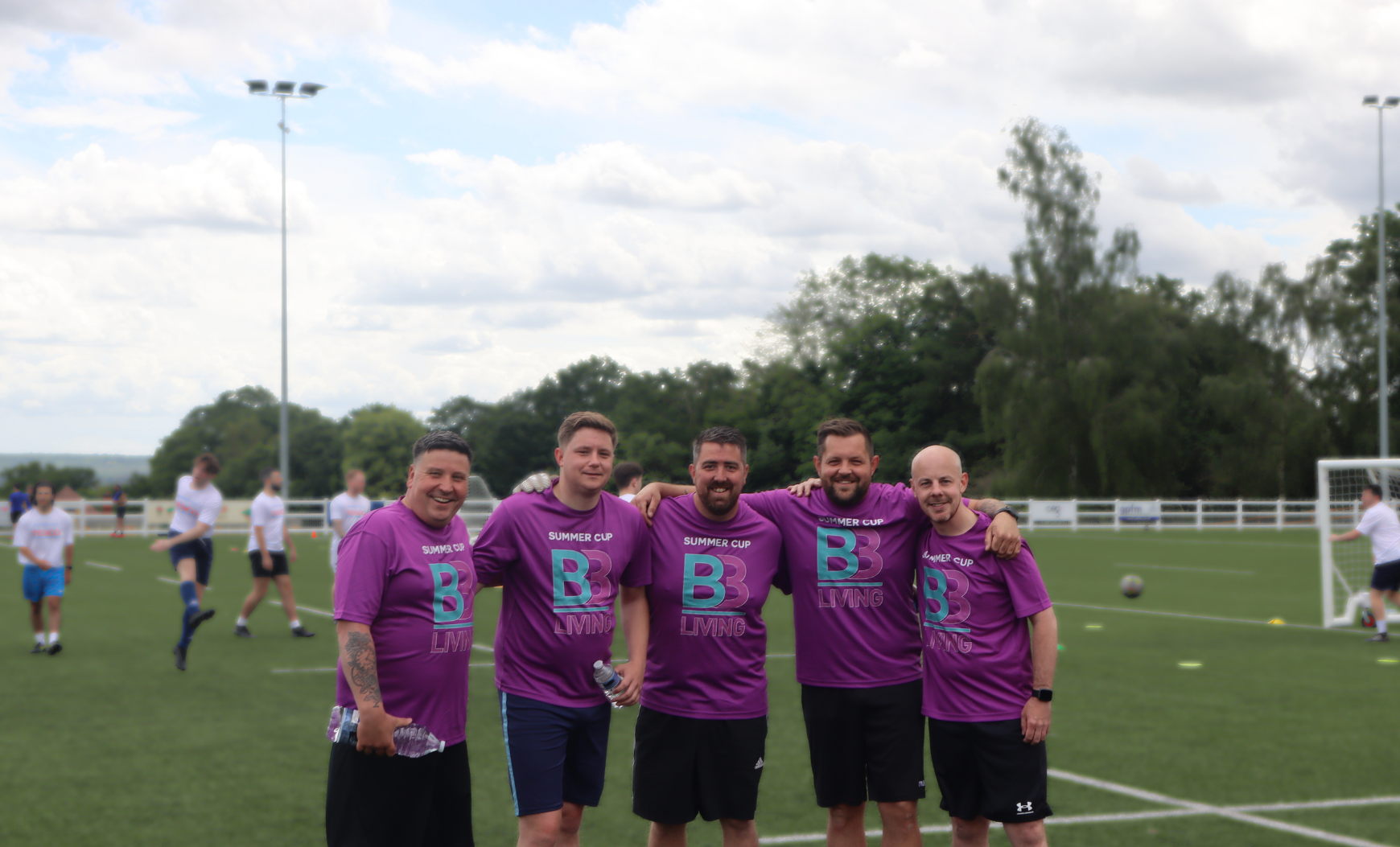 B3Living football team standing together in a football pitch. In the background, there are other players and a goalkeeper jumping to catch a ball