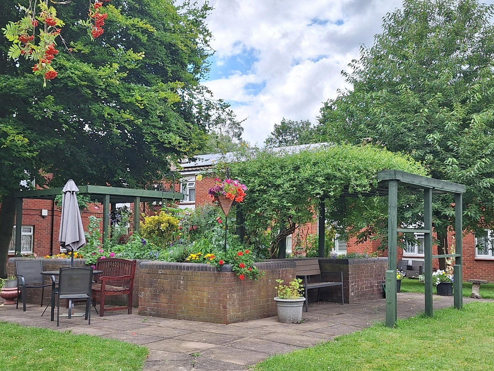 Garden patio with a table and chairs on the far left and a pergola on the right. There are trees on the sides and a square with colourful flowers and plants in the middle.