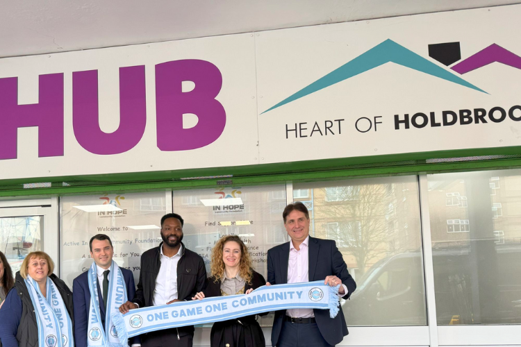 Five people including B3Living's CEO Steve Woodcock, Lewis Cocking MP, Mayor Sherrie McDaid and Patrick Kanyuka posing in front of the Holdbrook Hub. Three of them are holding a scarf that says 'One game one community' and the other two are wearing them on their necks.