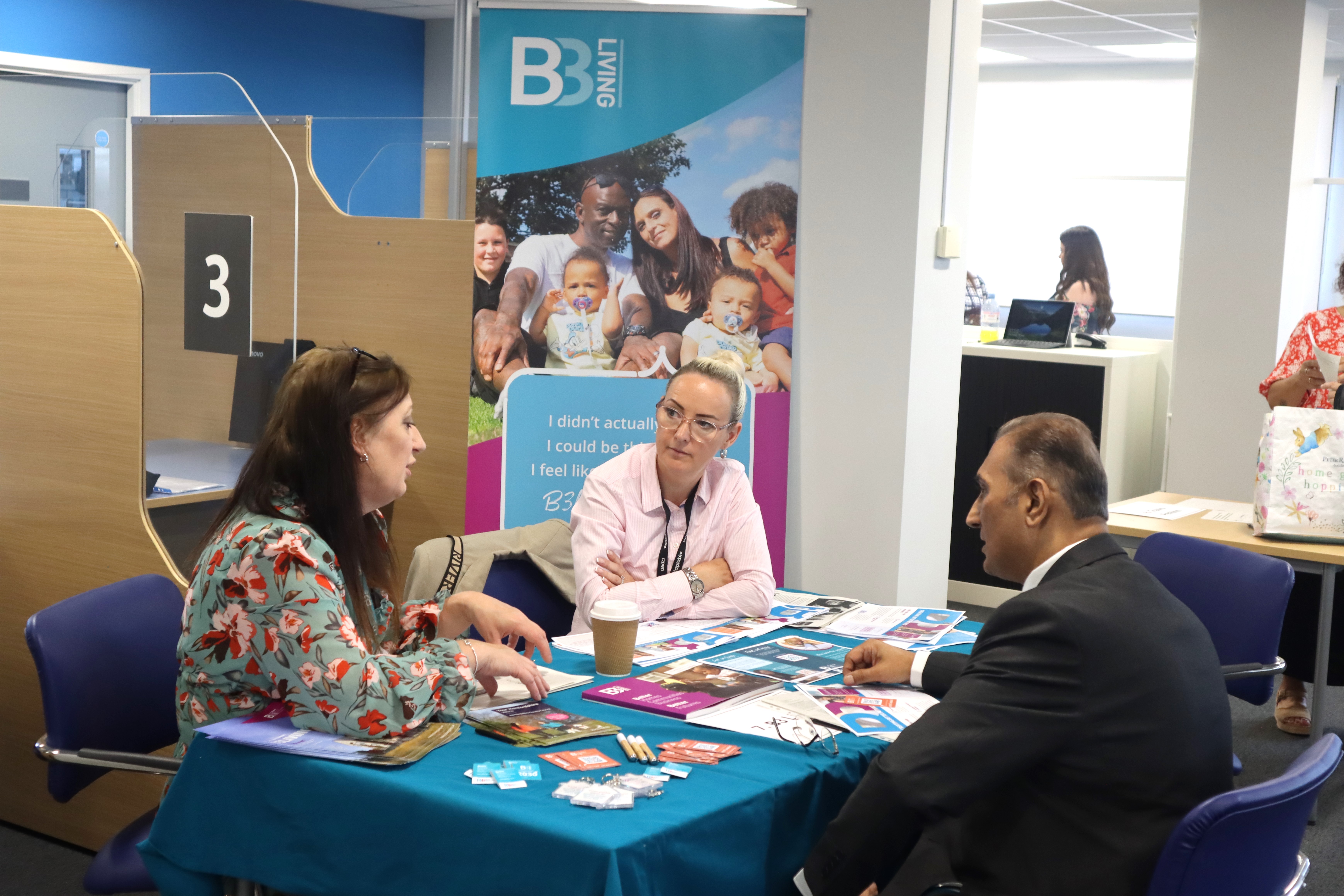 A man sitting across two B3Living female colleagues. They are talking over a table full of leaflets and booklets. Behind the two colleagues, there is a B3Living pull-up banner showcasing the brand logo, an image of a family of six which has some writing underneath that is covered by one of the colleagues.