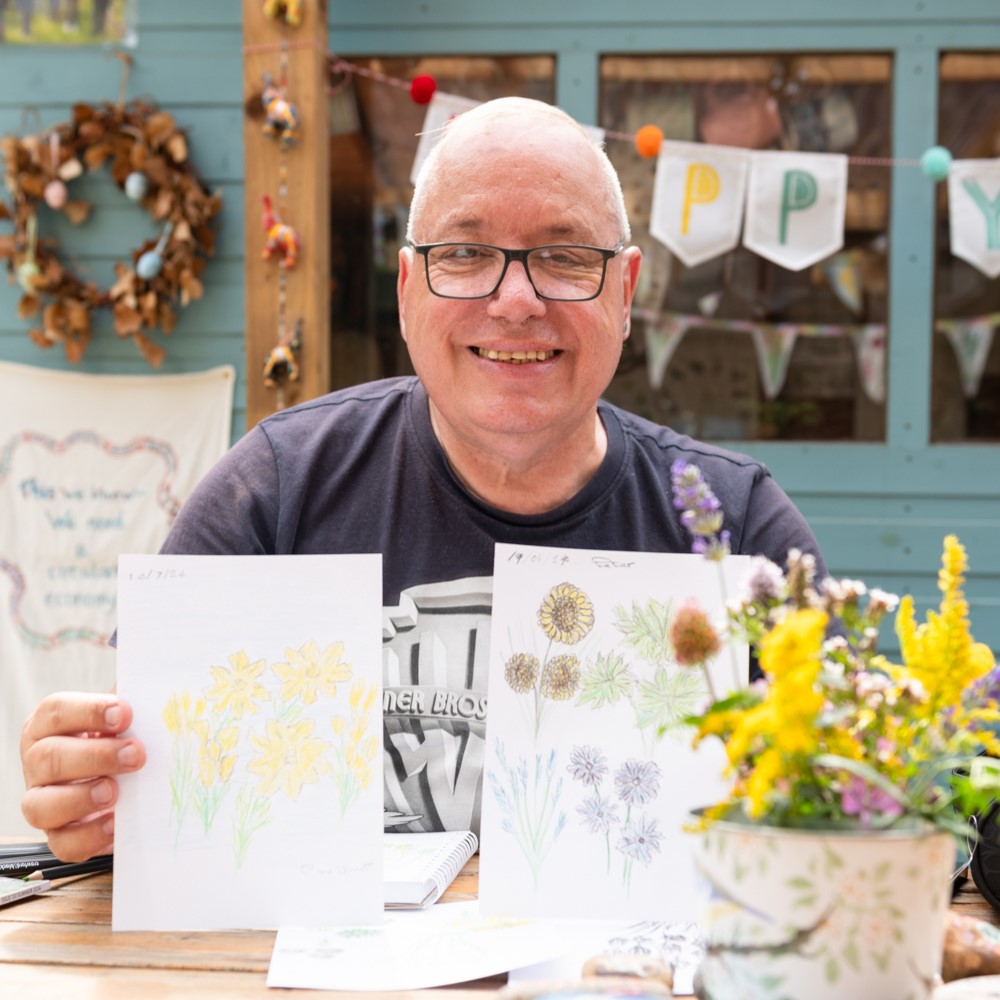 Peter smiling and holding up two drawings of flowers. There is a pot of yellow flowers slightly in front of the right side drawing