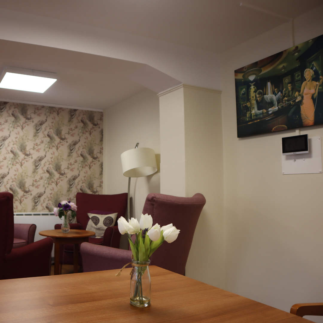 A living room with a table with a vase of white tulips on top. In the back, there are four sofa chairs facing each other and a lamp. On the right side, there is a wall-mounted Appello alarm.