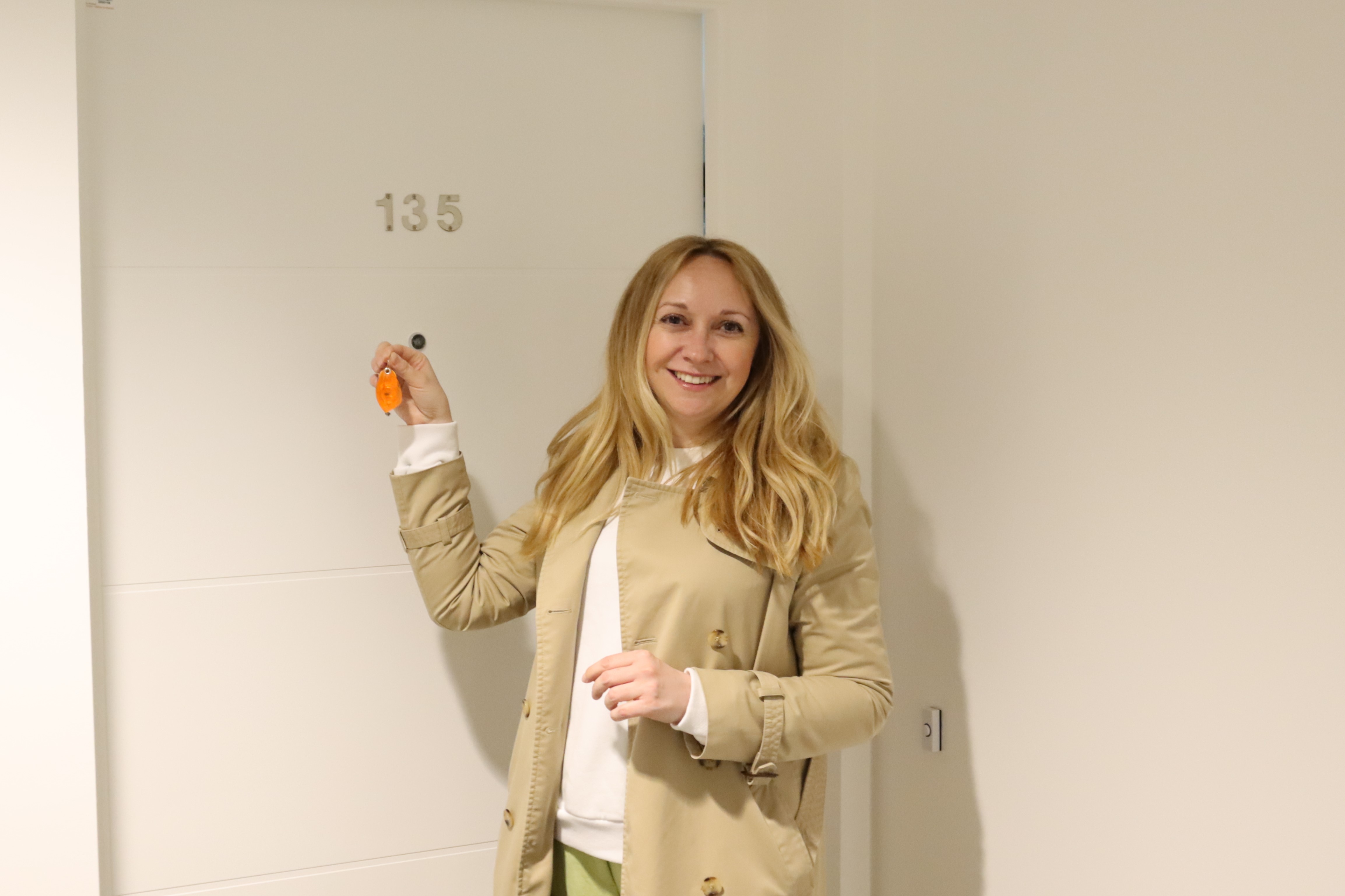Blonde, white woman smiling in front of a door numbered 135. She is wearing a beige trench coat.